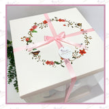 Top-down view of the gift box lid featuring a floral wreath and bird design, finished with a soft pink ribbon.