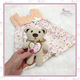 Hand holding a small teddy bear with a floral heart design, against a floral fabric background.