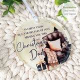 Close-up of the acrylic “Empty Chair” remembrance bauble personalised with Dad, showing the chair, tree and gift artwork on a cream knitted background with greenery.