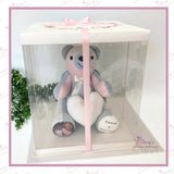 Front view of the clear plastic gift box showing the bear through transparent sides, with white base and pink ribbon tied at the top.