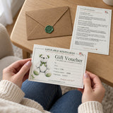 Held in hands: “Hands holding a printed gift voucher, with the matching information card and kraft envelope on a wooden table.