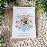 A positivity flower greeting card with a colorful flower illustration and supportive words, placed on top of a brown envelope over a knitted cream blanket with green leaves around the edges.