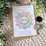 A floral positivity greeting card and kraft envelope laid on a knitted cream blanket, surrounded by green leaves and small flower embellishments. The card reads 'A Little Positivity Flower Just For You' with a central flower graphic containing words like 'You are loved,' 'You are strong,' 'You are special,' and 'You are enough.