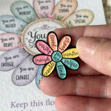 A hand holds a small flower-shaped enamel pin with colorful petals and positive self-affirmations, in front of a larger matching card with similar text.