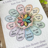 A close-up of a printed card or art print featuring a flower-shaped layout with pastel petals and centered text reading 'You are [adjective]' around a multicolored heart. The print is photographed on a neutral surface with a small leaf at the top right edge.