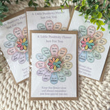 Handmade positivity flower cards arranged on a textured cream knit blanket with green leaves in the background. The central card is fully visible, showing a colorful flower made of pastel petals with positive affirmations like 'You are loved' and 'You are enough'.