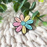 A flower-shaped enamel pin with multicolored petals and black outlines, featuring positive affirmations like 'loved', 'worthy', 'enough', 'capable', 'unique', and 'kind' on a chunky cream knit background with green leaves.