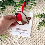 Hand holding a felt Christmas robin hanging decoration presented on a linen-textured remembrance card with the words “Robins appear when loved ones are near.