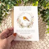 A6 robin remembrance keepsake poem card held by hand to show its size, with the removable enamel pin displayed inside an autumn woodland wreath.