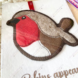 Single felt robin decoration laid flat on its remembrance card, showing embroidery details and red ribbon for hanging.